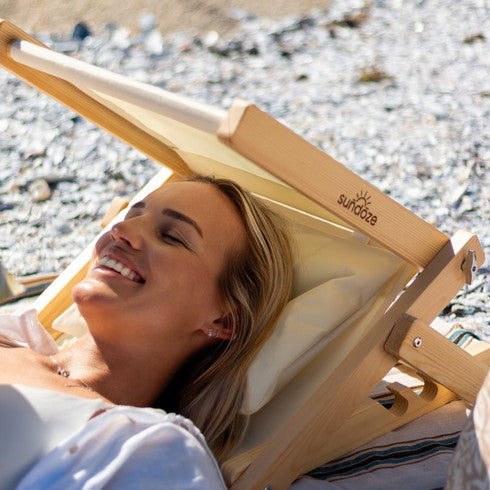 A woman smiling while resting her head  on a wooden folding SunDoze beach headrest with cream fabric on a pebble beach. 