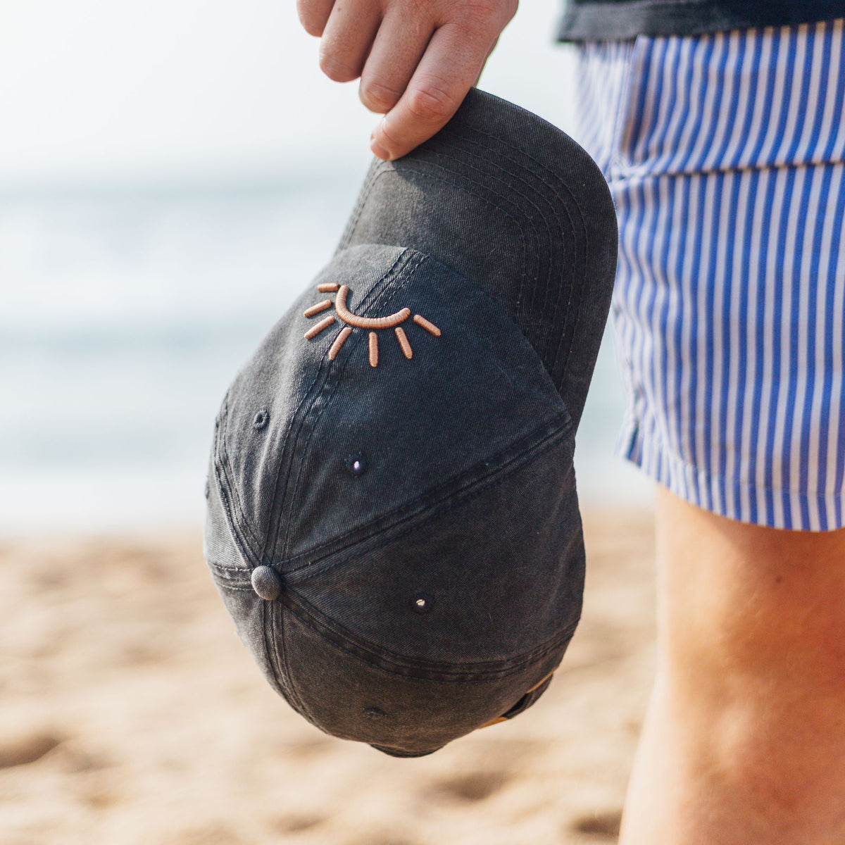 A person on a beach holds a washed black SunDoze cap.