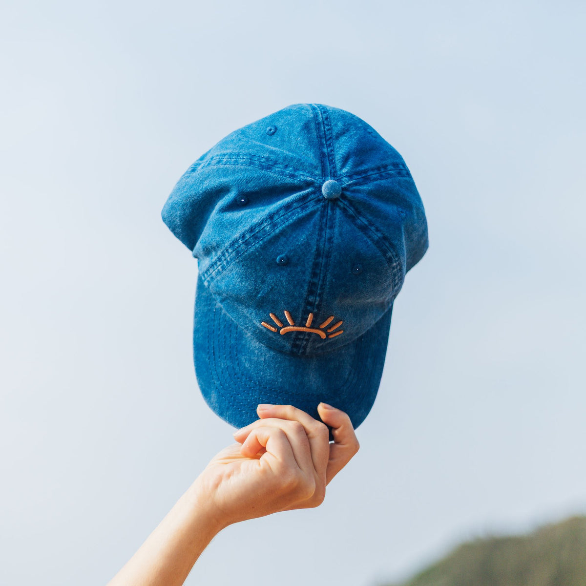 A blue SunDoze cap held up by a hand against a pale sky background.