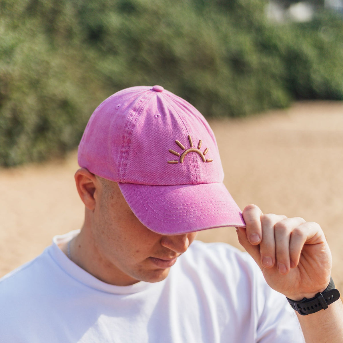 A man wearing a pink SunDoze cap, holding the brim down with his hand in an outdoor setting.