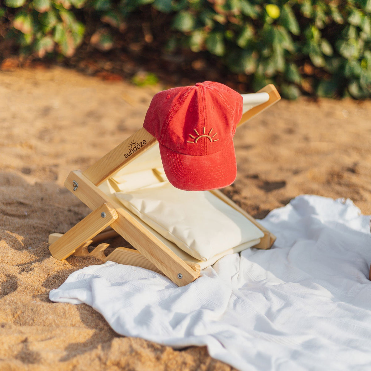 A red SunDoze cap rest on a SunDoze beach headrest  set on a white blanket on sandy ground with green foliage in the background.